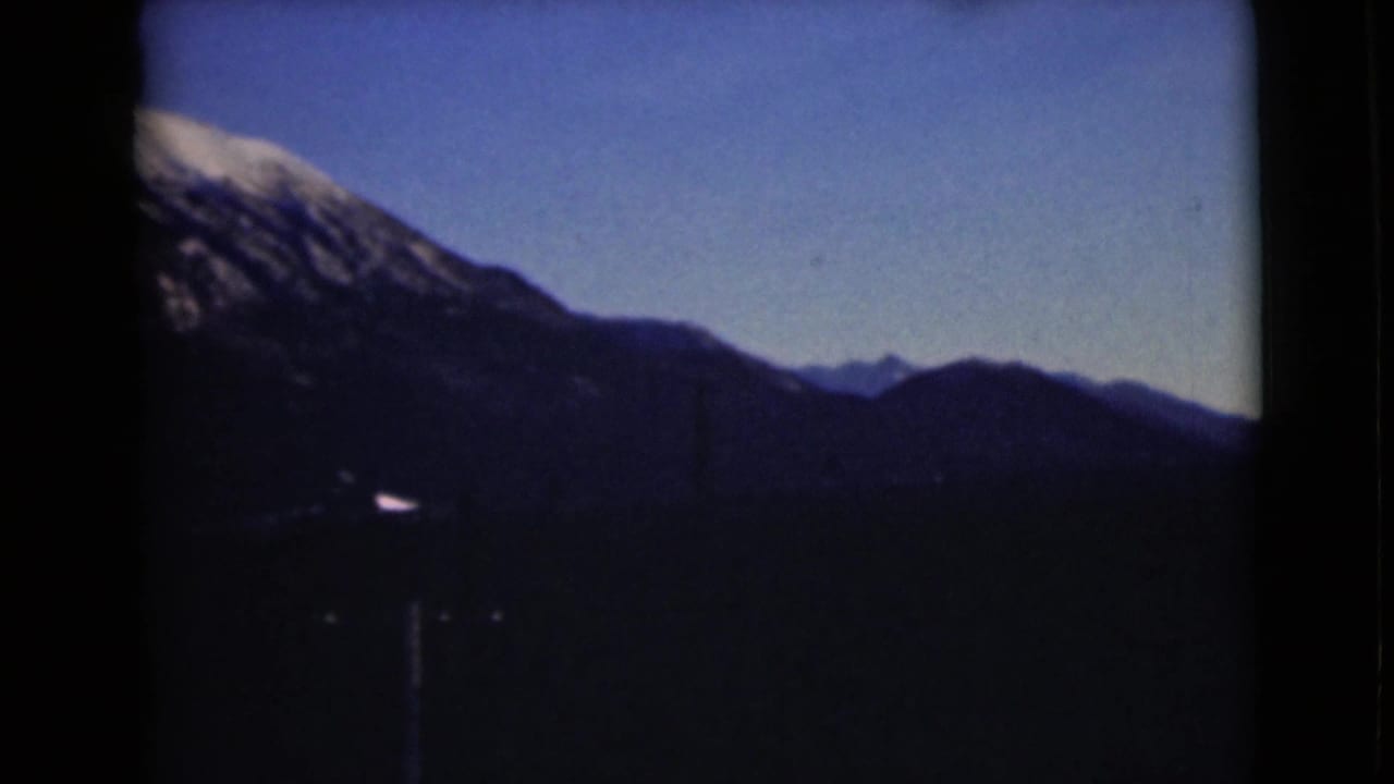 1946 Alaska 8mm view of a snow-capped mountain ridge above dark forested slopes, framed by rounded home-movie film edges.
