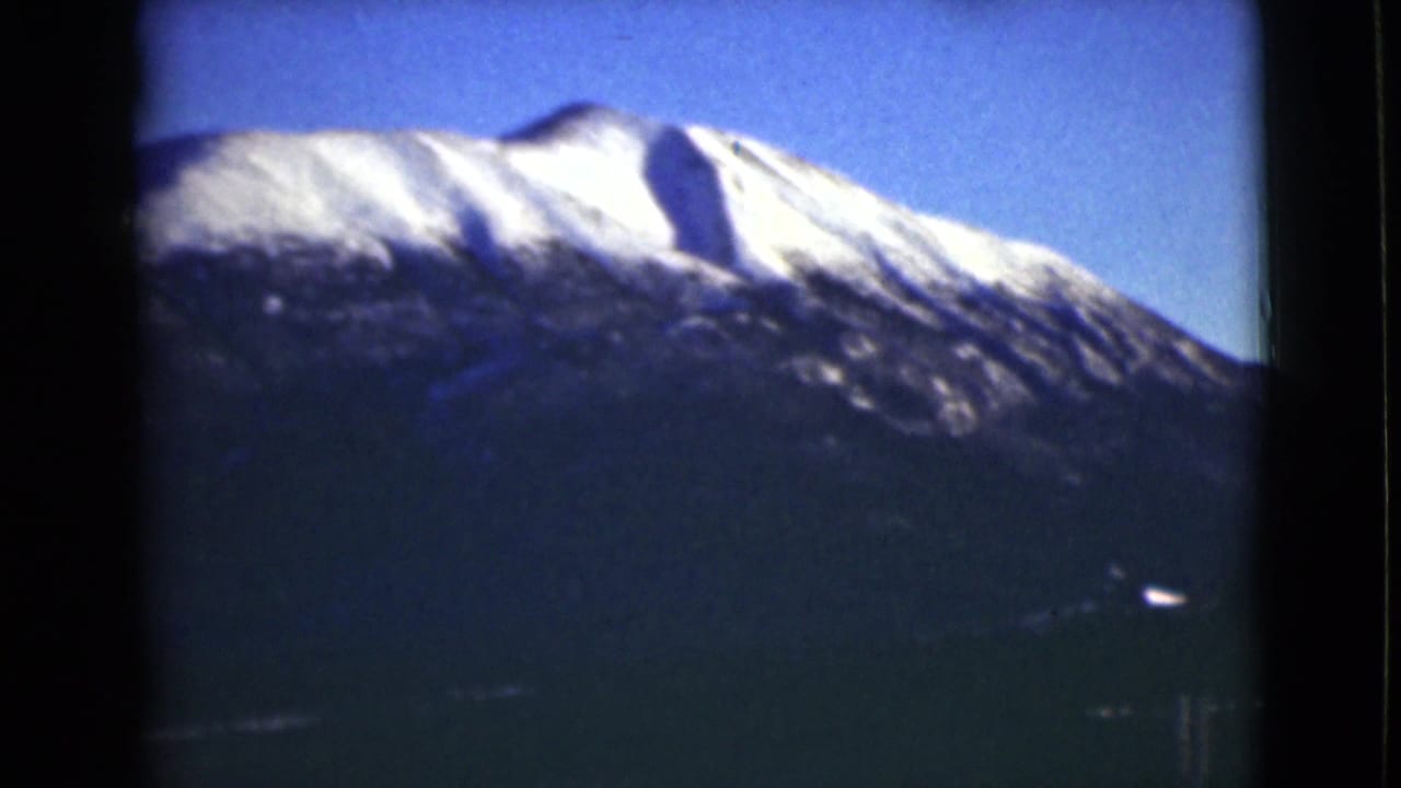 1946 Alaska 8mm frame showing a snowy mountain summit rising above a dark forested slope beneath clear blue sky.