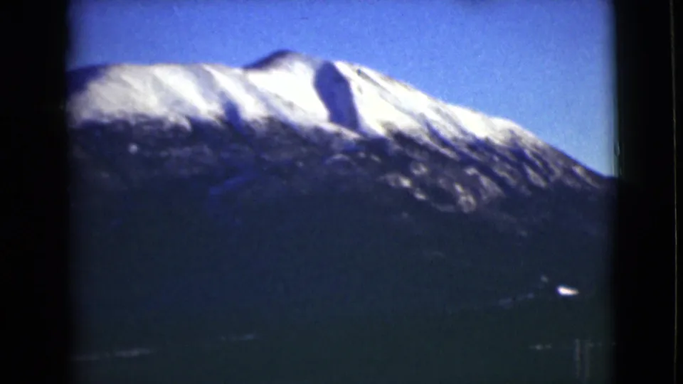 1946 Alaska 8mm frame showing a snowy mountain summit rising above a dark forested slope beneath clear blue sky.