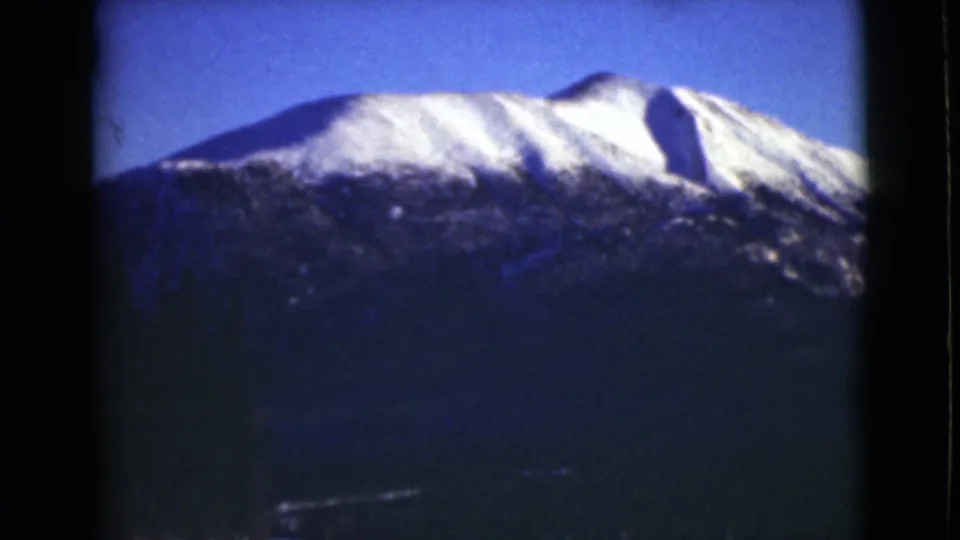 1946 Alaska home-movie frame showing a snow-bright summit above shadowed mountain terrain and a clear blue sky.