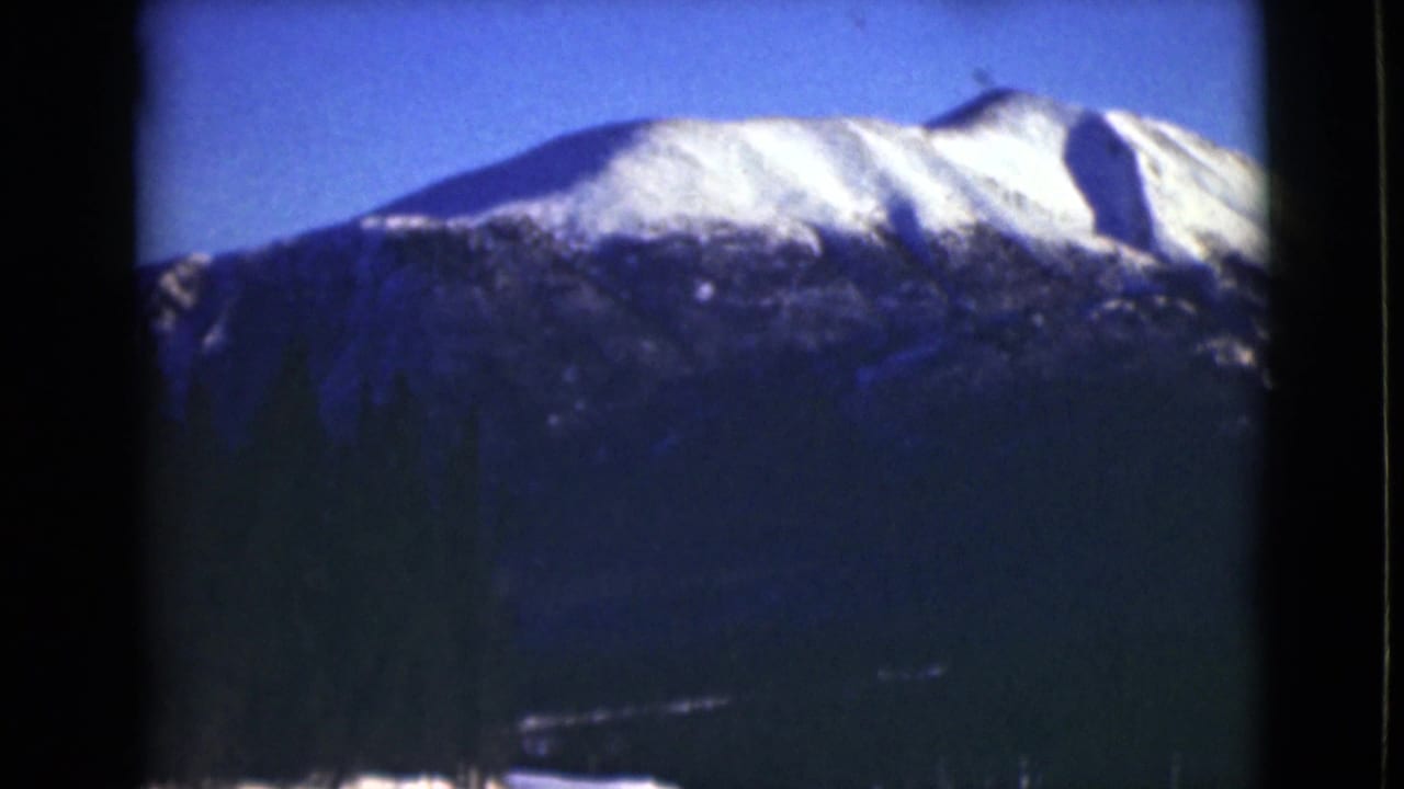 1946 Alaska 8mm frame showing a distant snow-capped mountain ridge beyond a dark evergreen forest and clear blue sky.