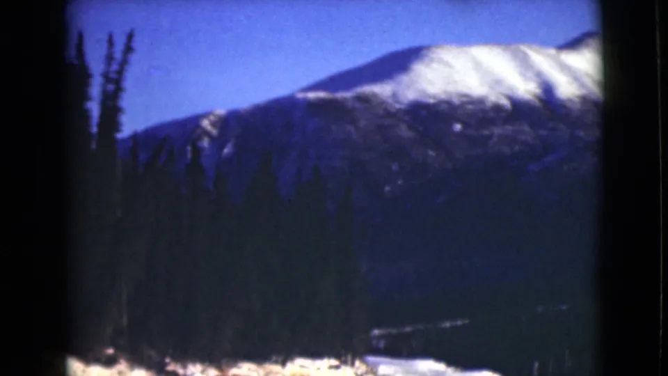 1946 Alaska 8mm frame with a snow-covered mountain slope, silhouetted evergreen trees, blue sky, and visible film-frame edges.
