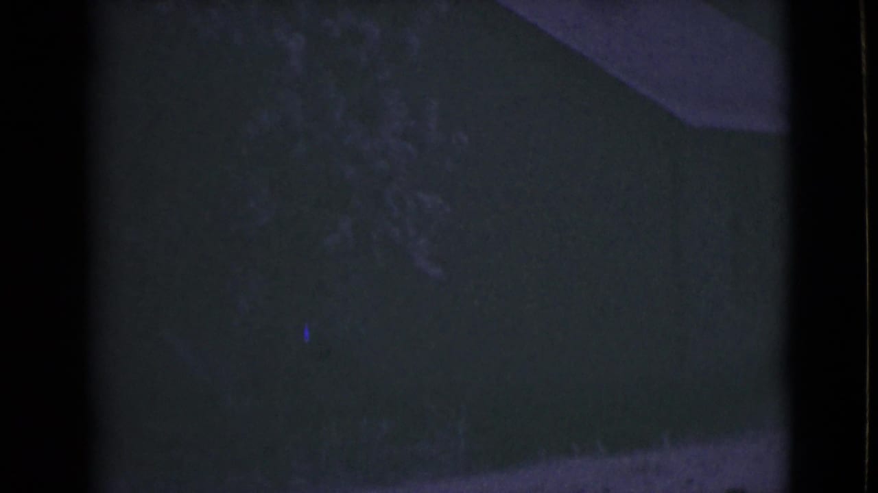 1946 Alaska home-movie frame showing a dim outdoor area with a shadowed building surface, vegetation, and a pale curb or path.