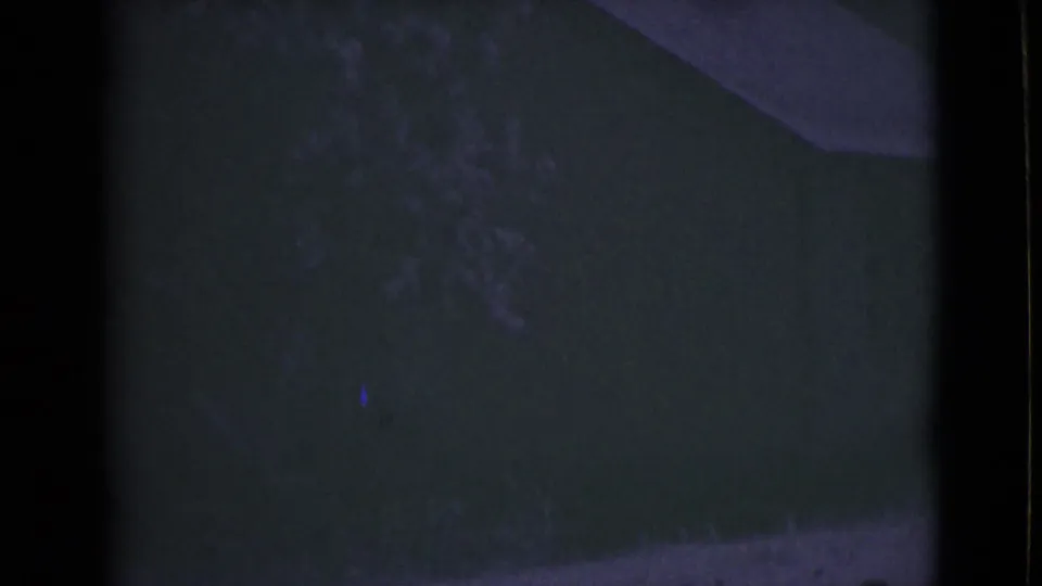1946 Alaska home-movie frame showing a dim outdoor area with a shadowed building surface, vegetation, and a pale curb or path.