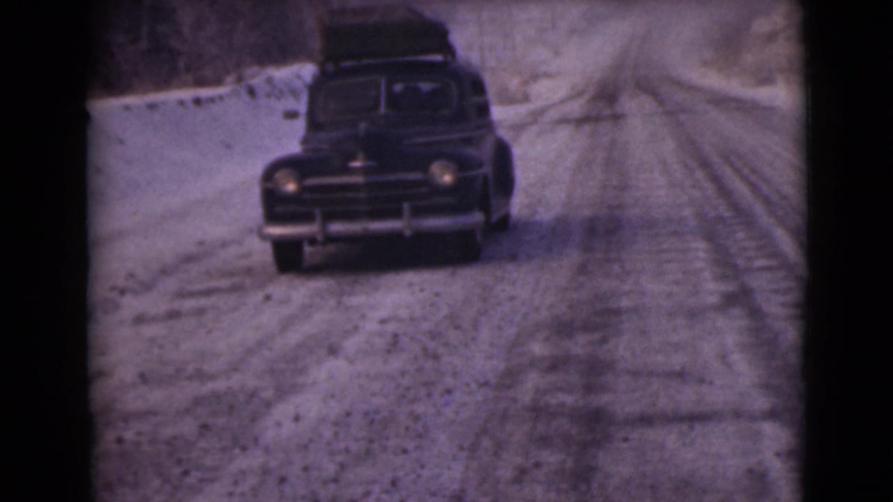 Dark vintage sedan with roof luggage driving toward the camera on a snowy road.