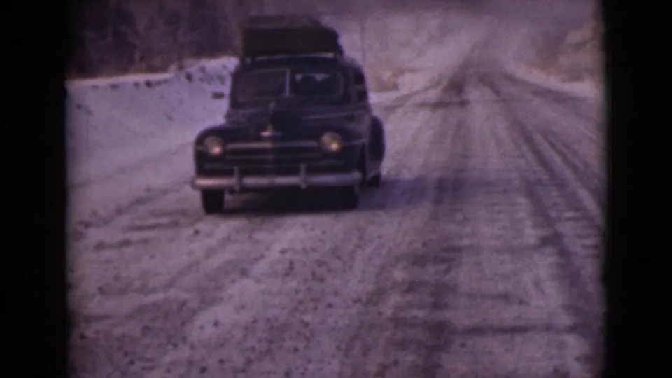 The car remains centered on the snowy Alaska road, with roof luggage and headlamps visible in the muted 8mm frame.