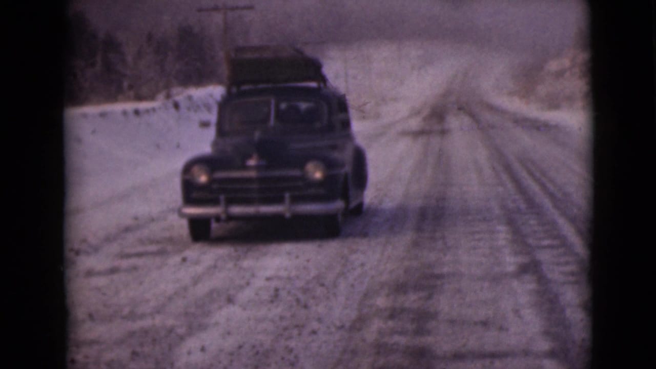 A black sedan with luggage on top approaches on a snow-dusted Alaska road in 1946 home-movie footage.