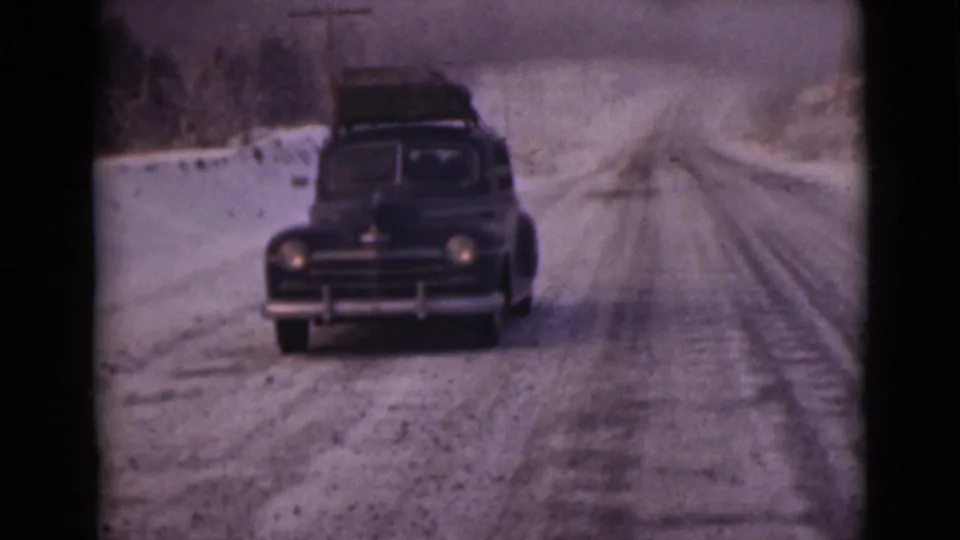 A black sedan with luggage on top approaches on a snow-dusted Alaska road in 1946 home-movie footage.