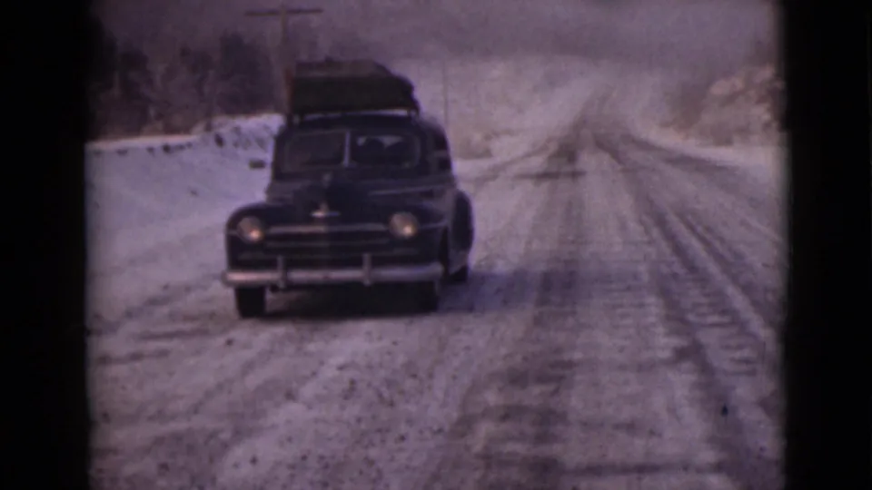The loaded black sedan continues toward the camera on a pale, snow-marked Alaska road.