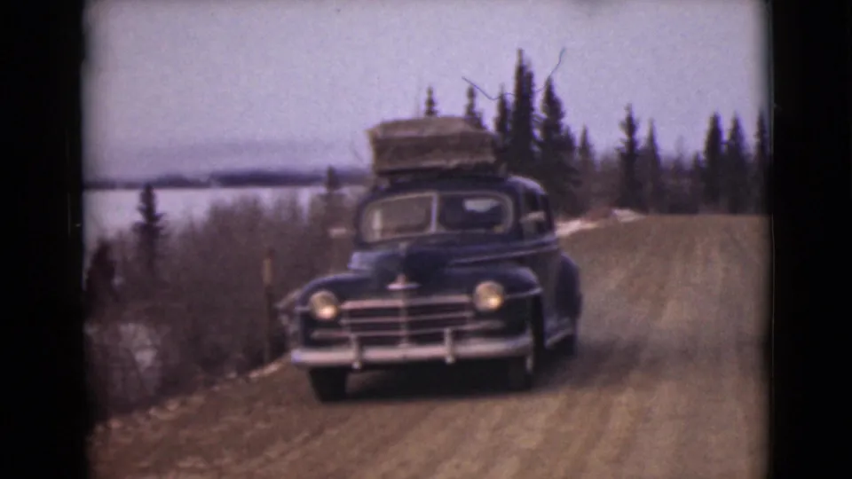 A loaded black sedan drives along a dirt road near pale water and evergreen trees in Alaska.