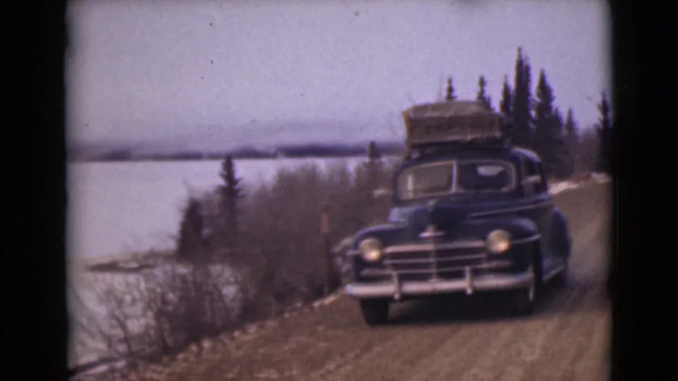 The black sedan passes close to the camera on a dirt road beside pale water and winter brush in Alaska.