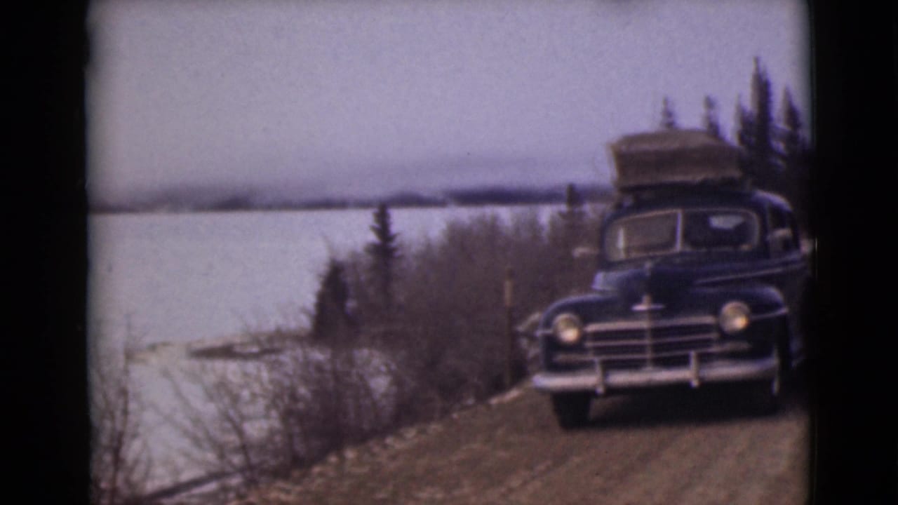 Dark vintage sedan with roof luggage parked on a dirt road beside a frozen lake.