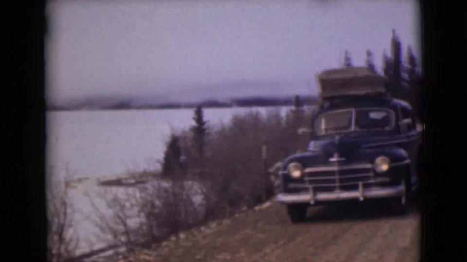 1946 Alaska home-movie frame showing a dark sedan with luggage on the roof near a frozen lake and sparse roadside trees.