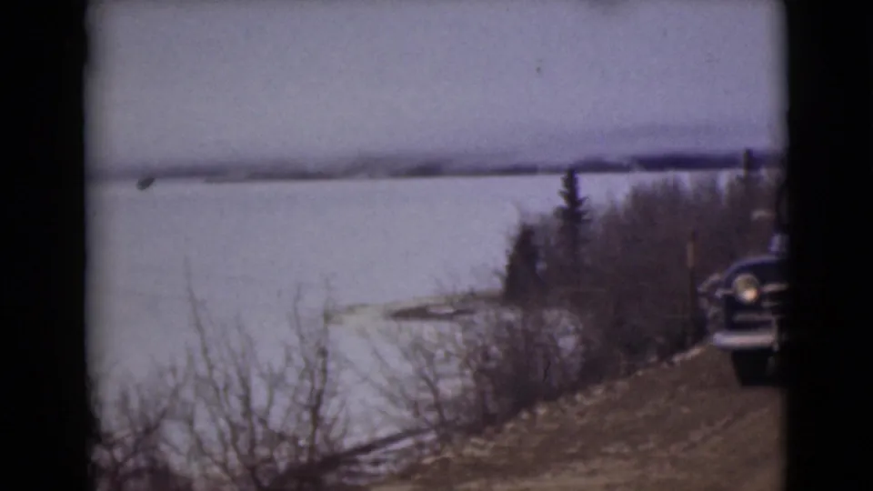 1946 Alaska roadside view with a frozen lake, brush, distant shoreline, and the edge of a dark sedan entering the frame.