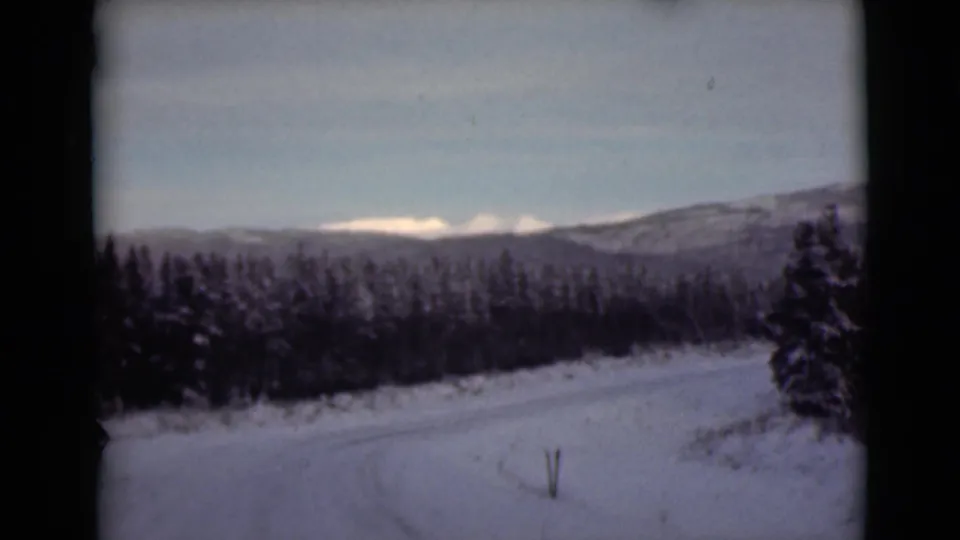 1946 Alaska 8mm frame showing a snow-covered road, dense conifers, pale sky, and distant mountain ridges.