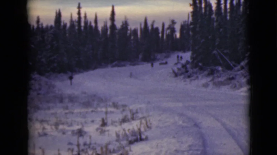 1946 Alaska 8mm frame of a snowy forest road or trail with faint tracks and small distant people in the landscape.
