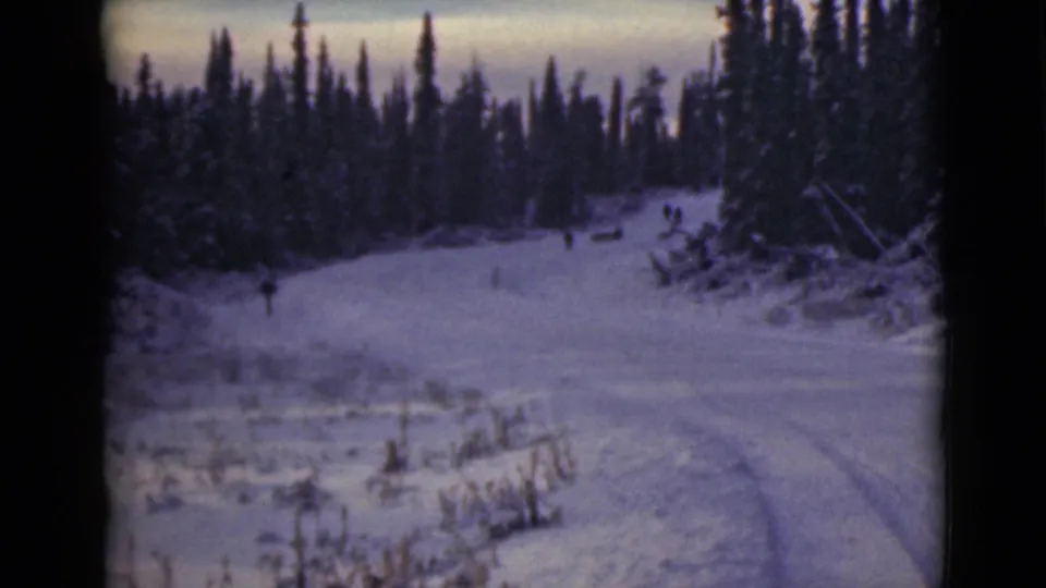 1946 Alaska home-movie frame of a curving snow path through evergreen woods, with tiny distant figures on the trail.