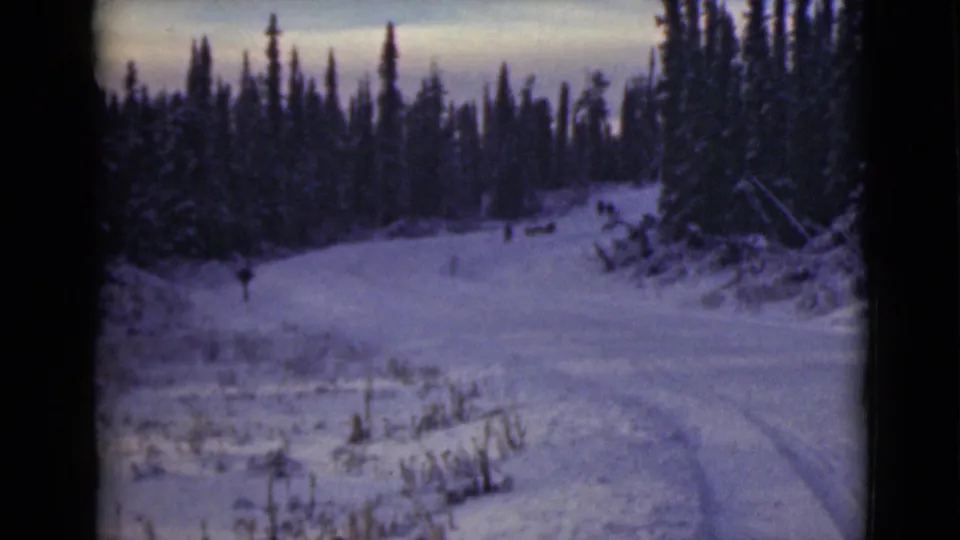 1946 Alaska 8mm frame of a snowy forest road with faint tracks and small distant people or animals near the bend.