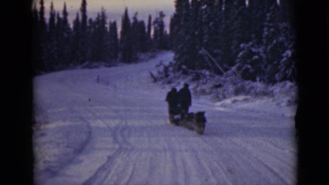 In Alaska archival 8mm footage, two bundled riders and a dog team move away on a snow-covered forest trail.