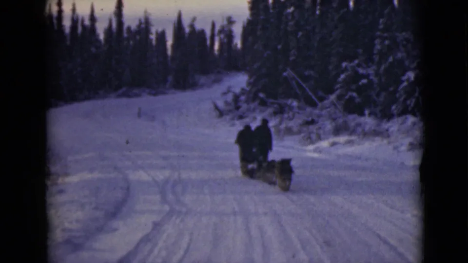 In Alaska archival 8mm footage, two bundled riders and a dog team move away on a snow-covered forest trail.