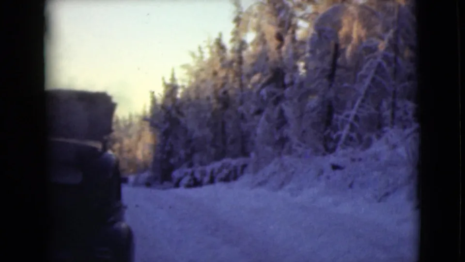 A dark vintage car appears at the edge of a snowy Alaska road, with frost-covered trees lining the route.