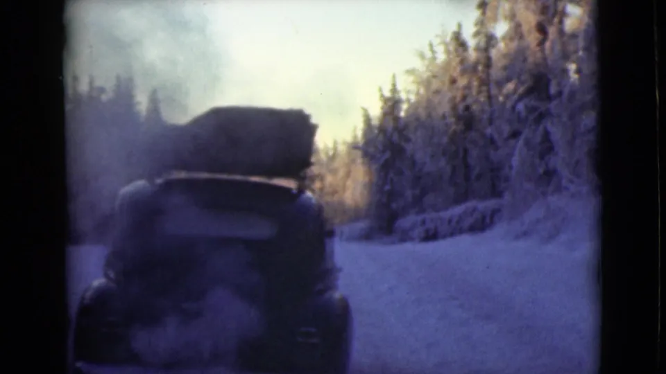 Rear view of a vintage car with roof cargo on a snowy Alaska road, with exhaust or cold vapor trailing in dusk light.
