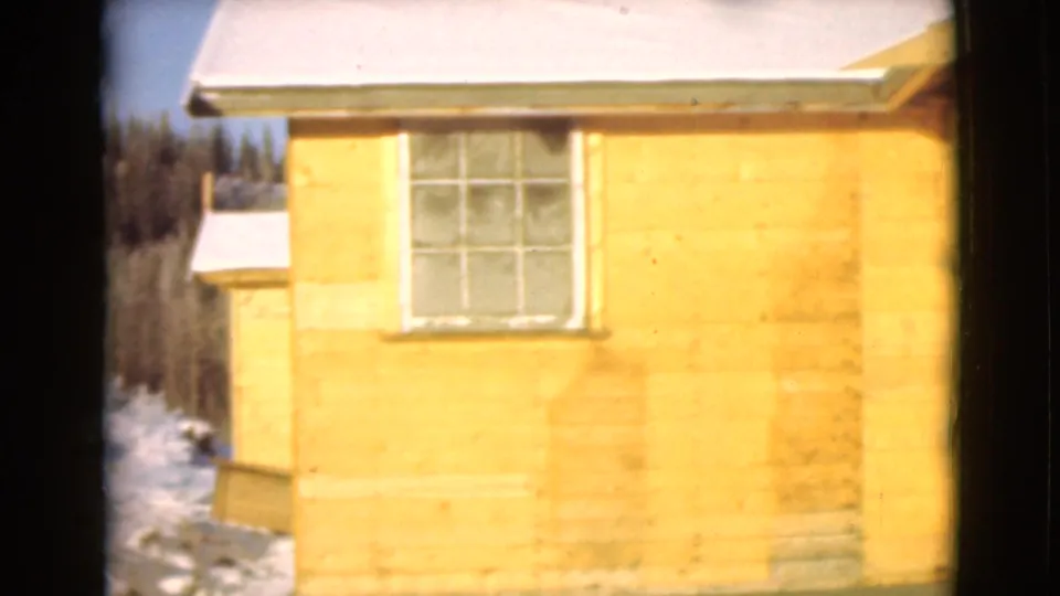1946 Alaska 8mm frame of a yellow wooden cabin exterior with a window, snow-dusted roof, and winter trees in back.