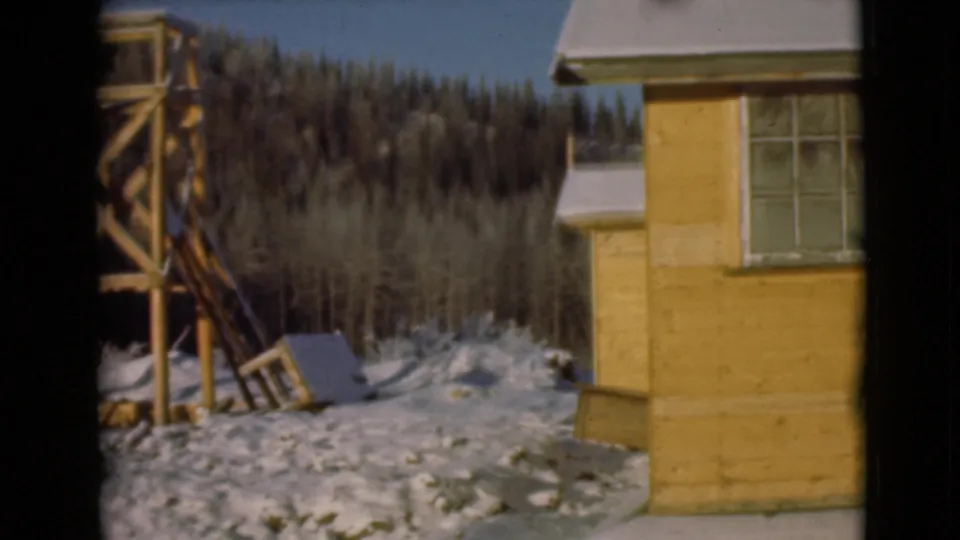1946 Alaska 8mm view of a yellow cabin side with a window, snowy ground, dark trees, and a nearby wooden raised structure.