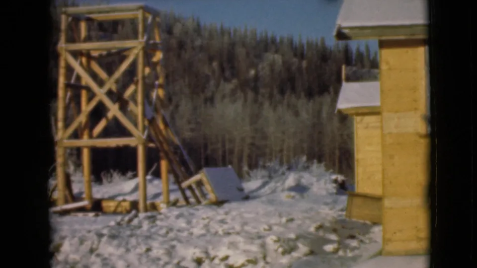1946 Alaska 8mm frame showing a timber watchtower, part of a cabin, snow-covered ground, and evergreen forest.