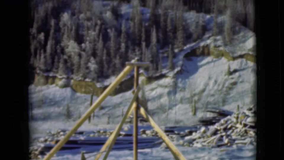 1946 Alaska home-movie frame with a wooden tripod-like structure, stacked timber, and snow-covered evergreen woods beyond.