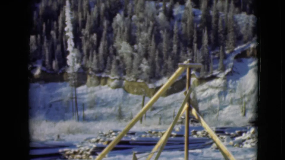 1946 Alaska 8mm frame showing a static view of a snowy clearing, tripod-like wooden frame, cut logs, and dark evergreen trees.