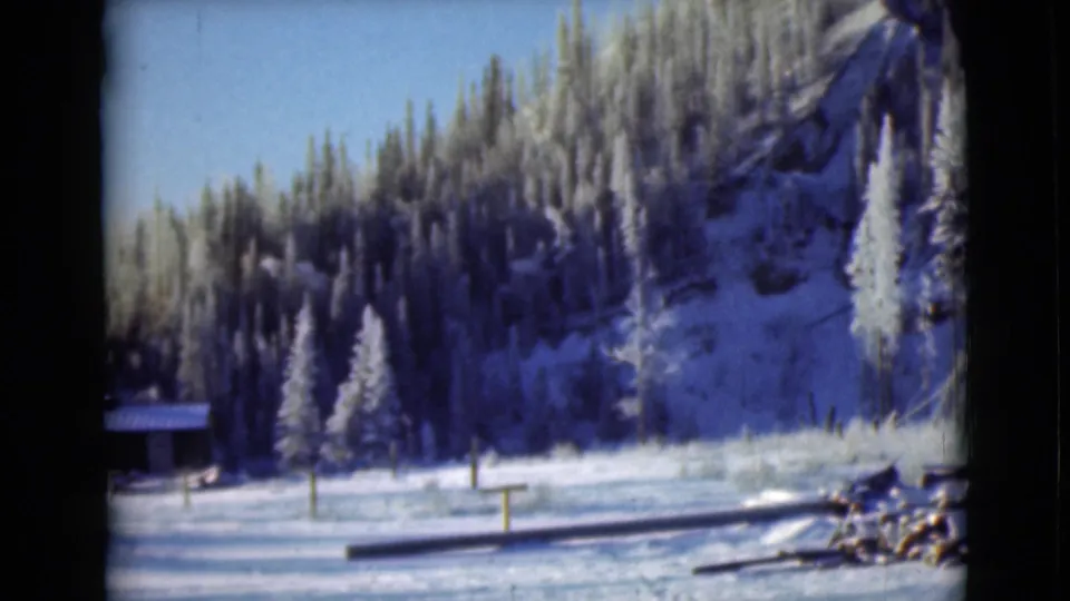 1946 Alaska 8mm view of a small wooden structure at the edge of a snowfield below a frost-covered evergreen slope.