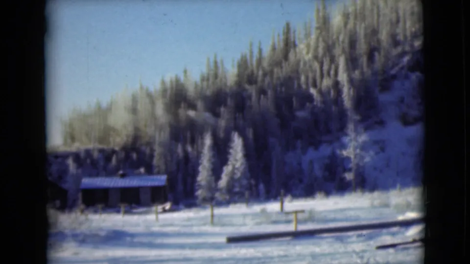1946 Alaska frame showing a small wooden building below a snowy evergreen slope, with posts and logs across the foreground.