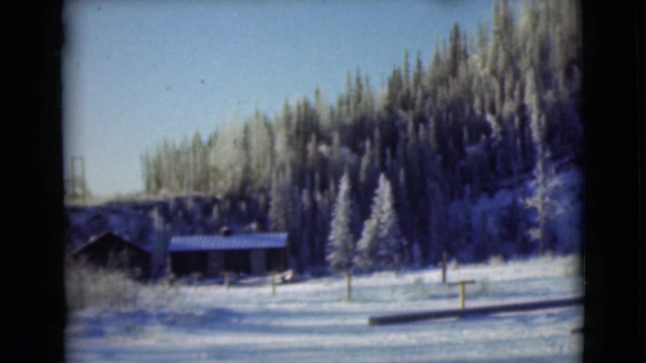 1946 Alaska home-movie frame showing snow-covered ground, dark cabin-like structures, and evergreen trees under a clear sky.
