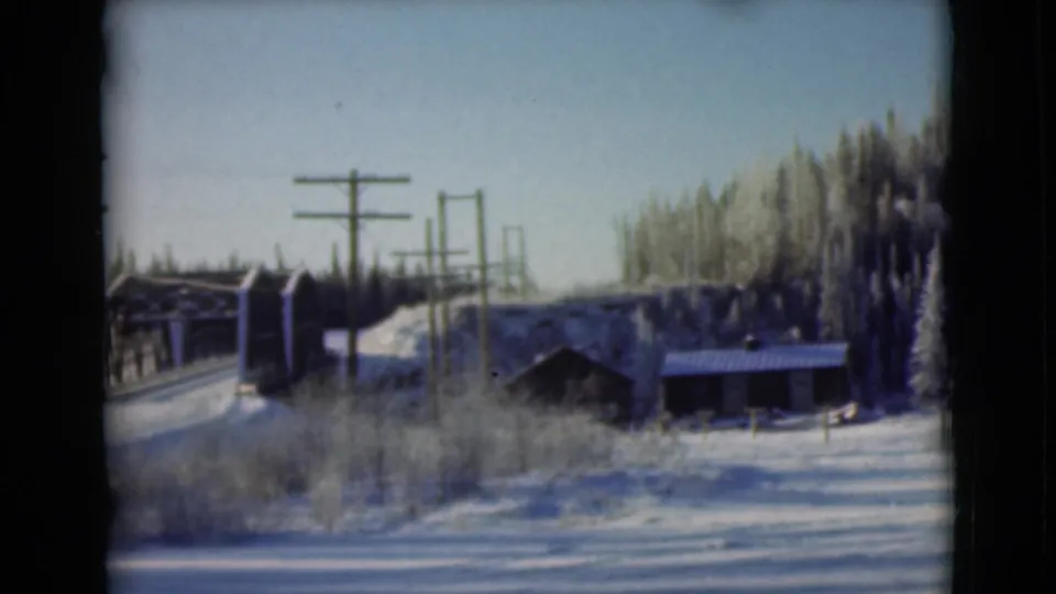 1946 Alaska frame showing a wooden bridge beside utility poles, snow-covered brush, a small structure, and winter evergreens.