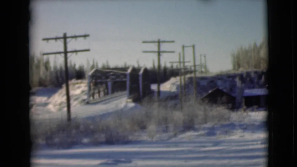1946 Alaska home-movie still of a snow-covered bridge area, utility poles, dark cabin-like structure, and forested hillside.