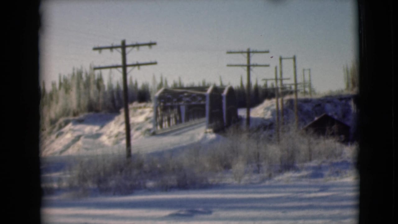 1946 Alaska 8mm frame showing a snow-covered roadside with wooden utility poles, winter brush, and a small bridge or trestle.
