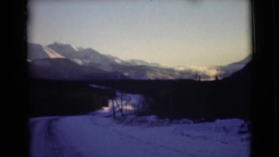1946 Alaska 8mm view of tire tracks curving through snow toward a dark tree line and distant mountain range.