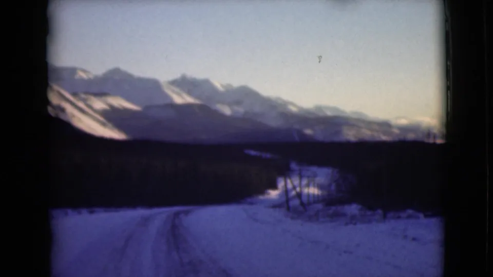 1946 Alaska winter frame showing a snow-covered road with tire tracks curving toward a distant mountain range under pale sky.