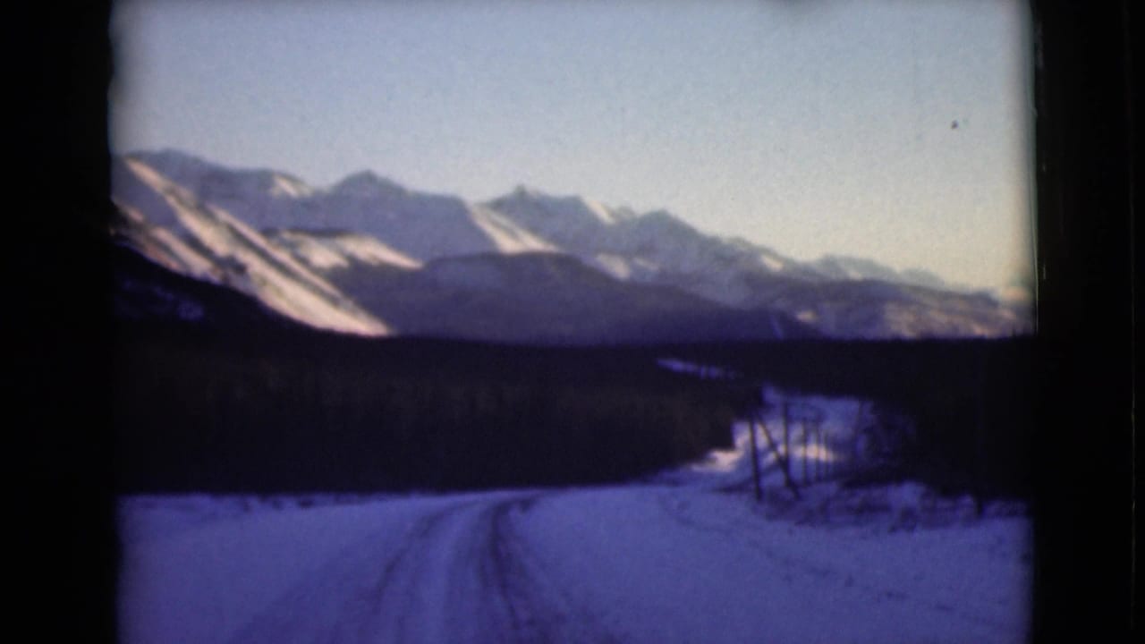 1946 Alaska 8mm frame showing a snow-covered road leading toward evergreen trees and distant mountain peaks under a pale sky.