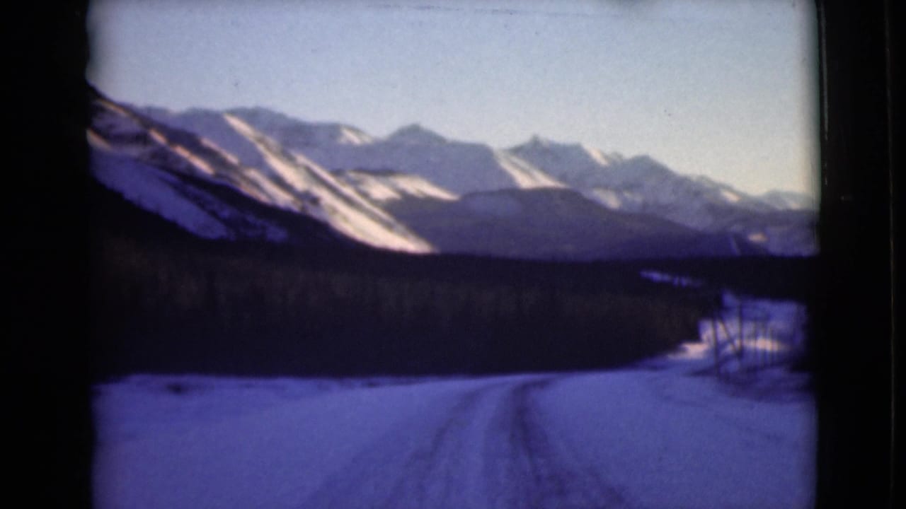 Winter road and dark tree line below a broad range of snowy peaks.