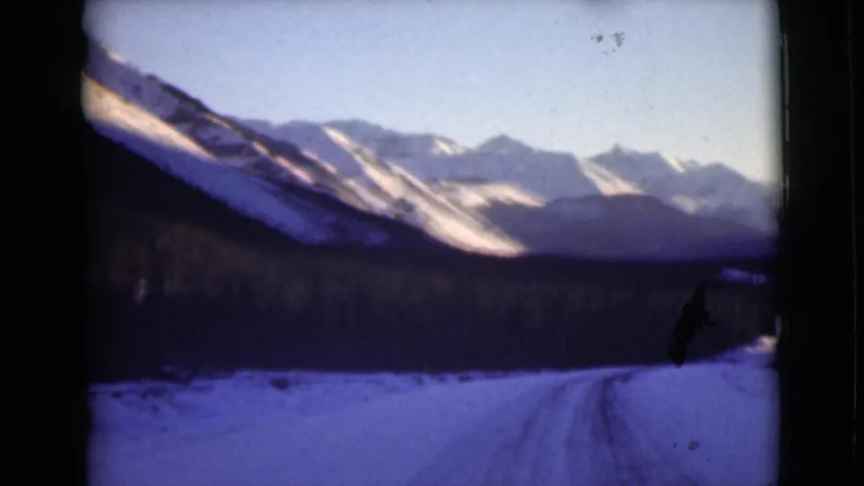 1946 Alaska 8mm frame showing a snowy roadside foreground, dark trees, and layered mountain ridges with sunlit snow patches.