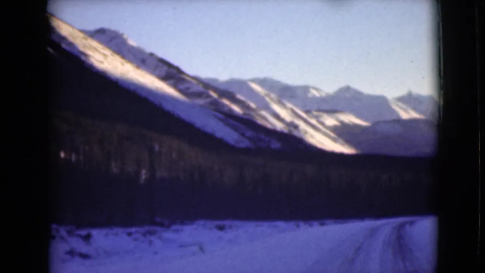 1946 Alaska home-movie frame with a road bending in from the right, a dark forest band, and snow-bright mountain slopes beyond.