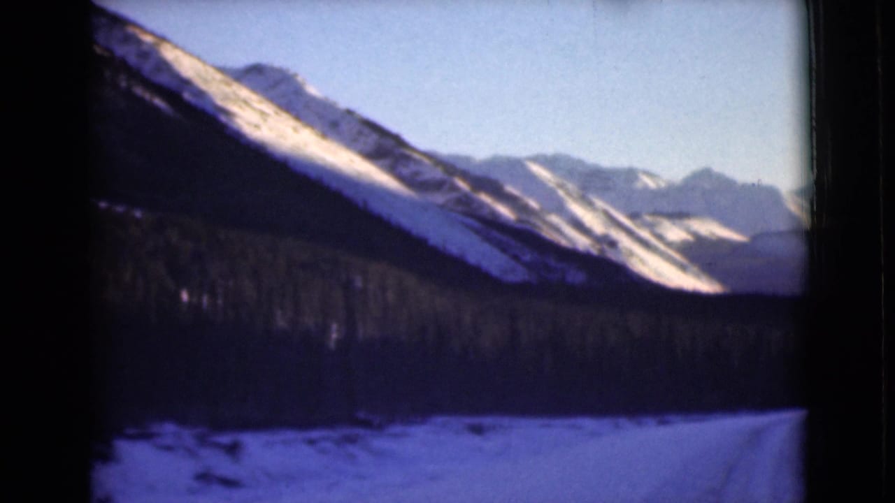 Snow-covered mountain valley with evergreen forest under a pale blue sky.