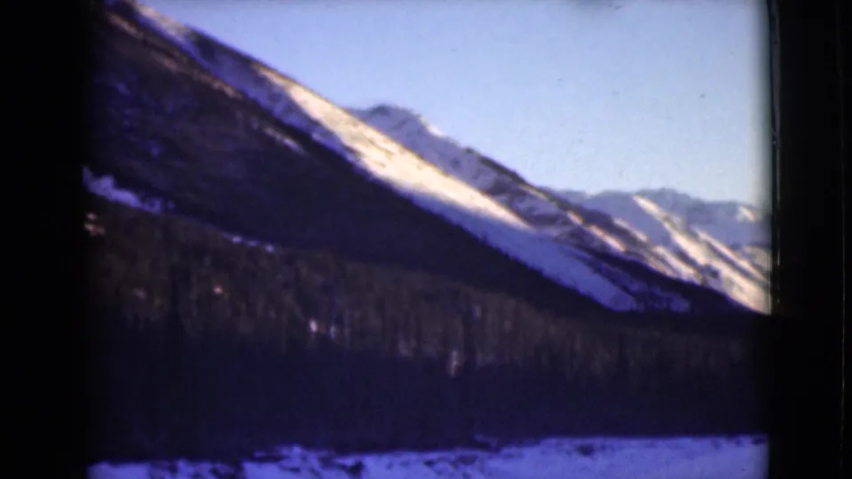 1946 Alaska 8mm frame with a steep snow-streaked mountain face, forest below, distant peaks, and soft blue sky.