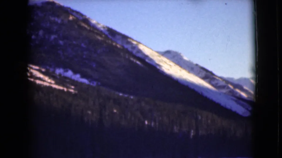 1946 Alaska 8mm frame showing a closer view of a snow-patched mountain slope, dark tree line, and clear sky.