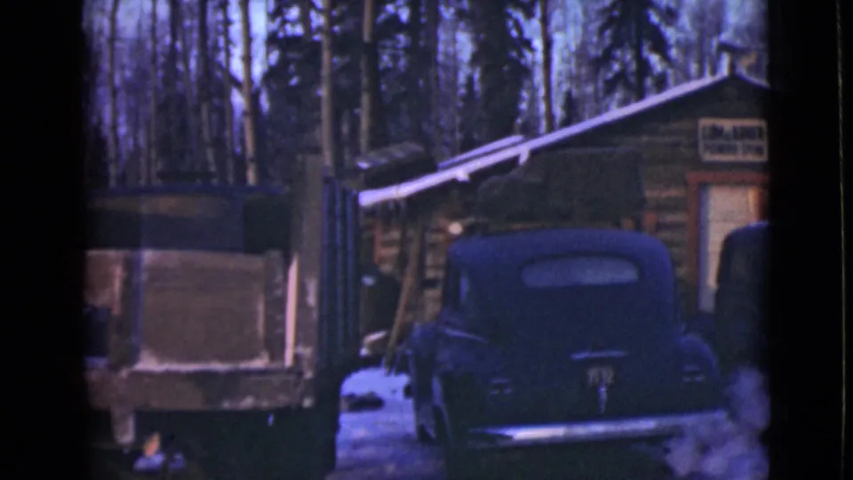 1940s Alaska home-movie frame showing parked automobiles, a truck or trailer, snow patches, trees, and a log building with a readable sign.
