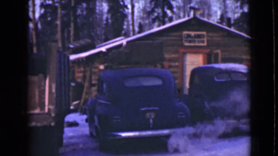 1940s Alaska 8mm frame of two rounded automobiles parked by a rustic log cabin, with snow on the ground and trees behind it.