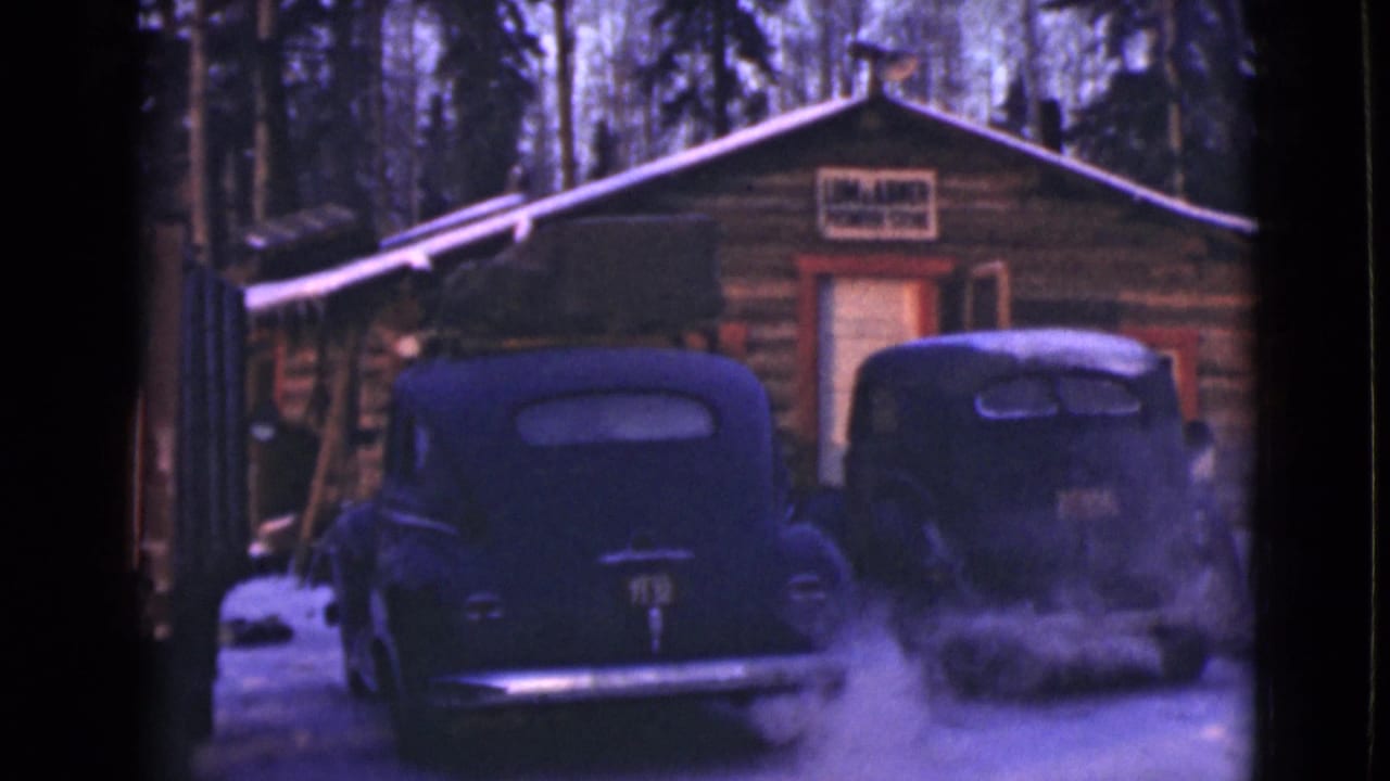 Two dark vintage cars parked in snow outside a log cabin with a sign over the door.
