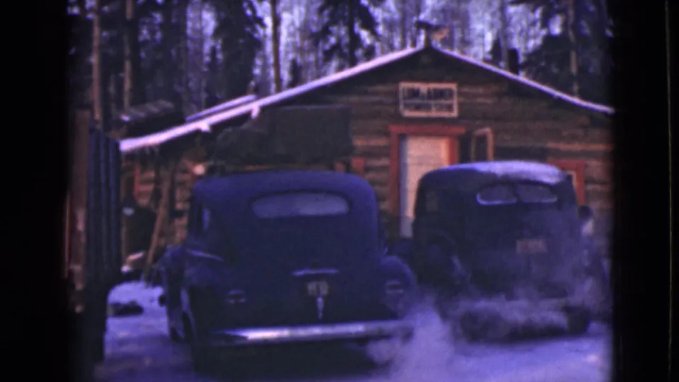 1946 Alaska 8mm frame showing two dark mid-century cars outside a snowy log cabin marked with a power station sign.